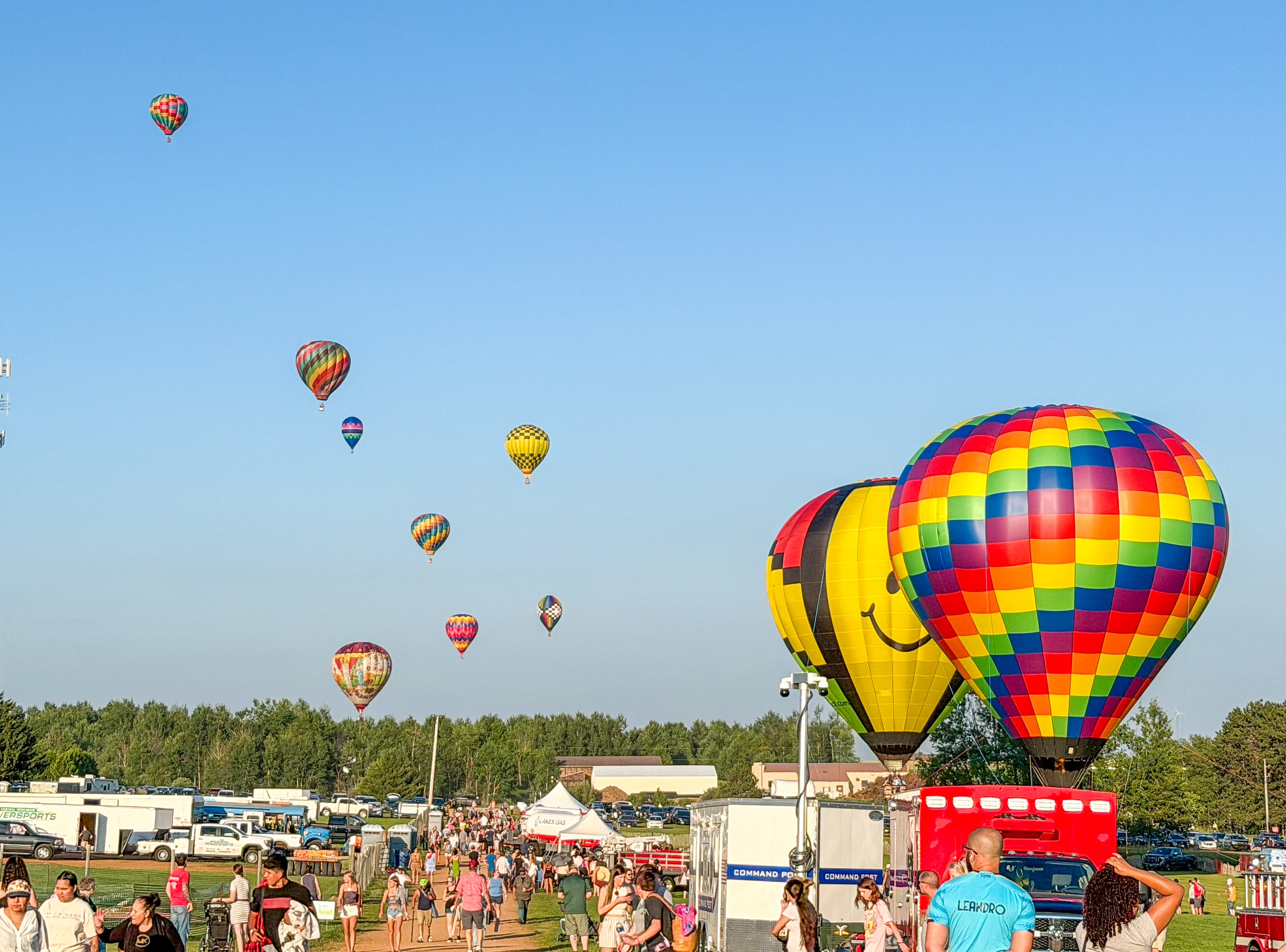 Vibrant hot air balloons float in a clear sky above a crowded outdoor festival and fairground.