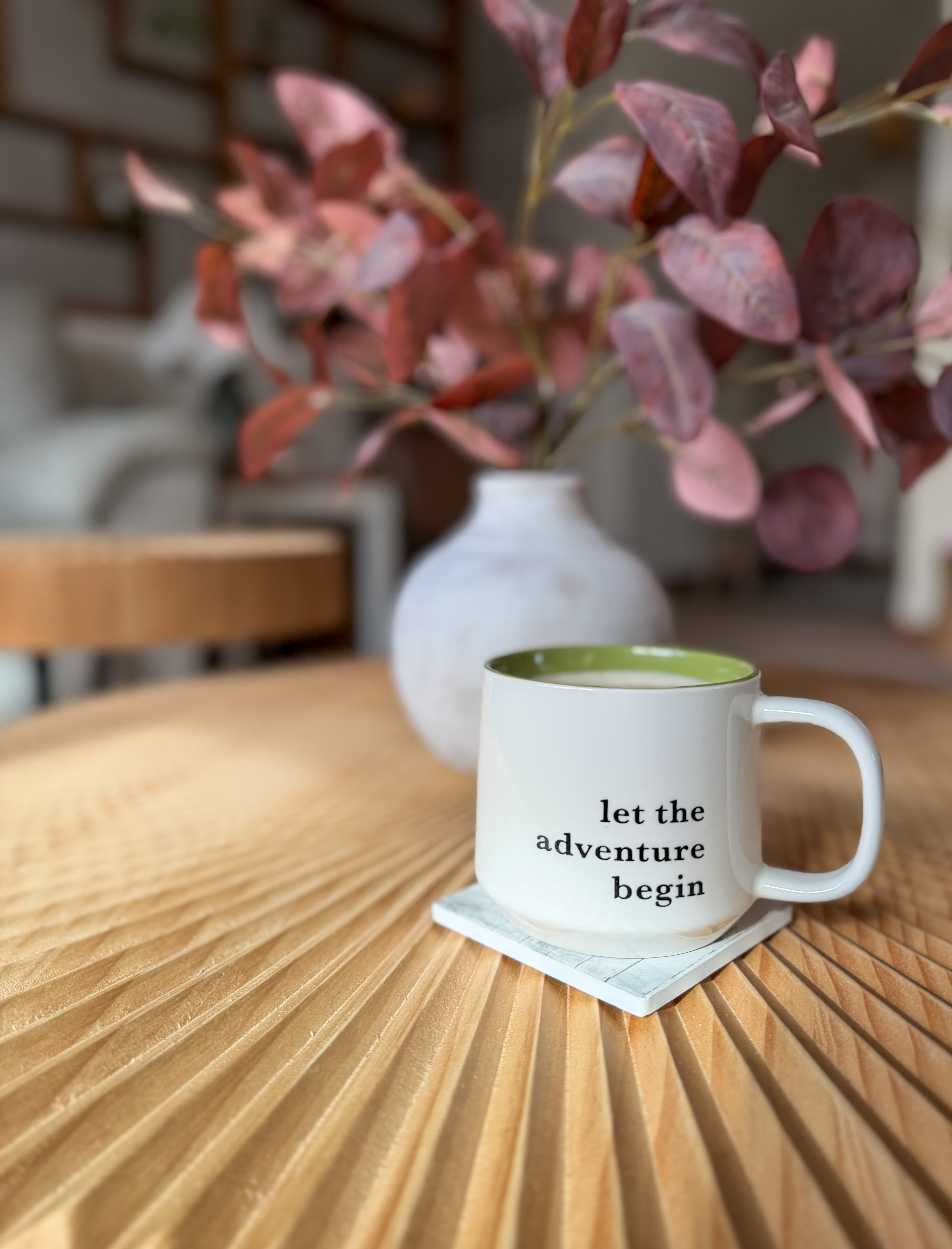 White mug reading 'let the adventure begin' on a fluted wooden table with autumn leaves.